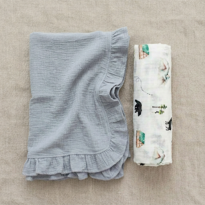 Blue ruffled baby blanket and a folded woodland swaddle on a beige surface